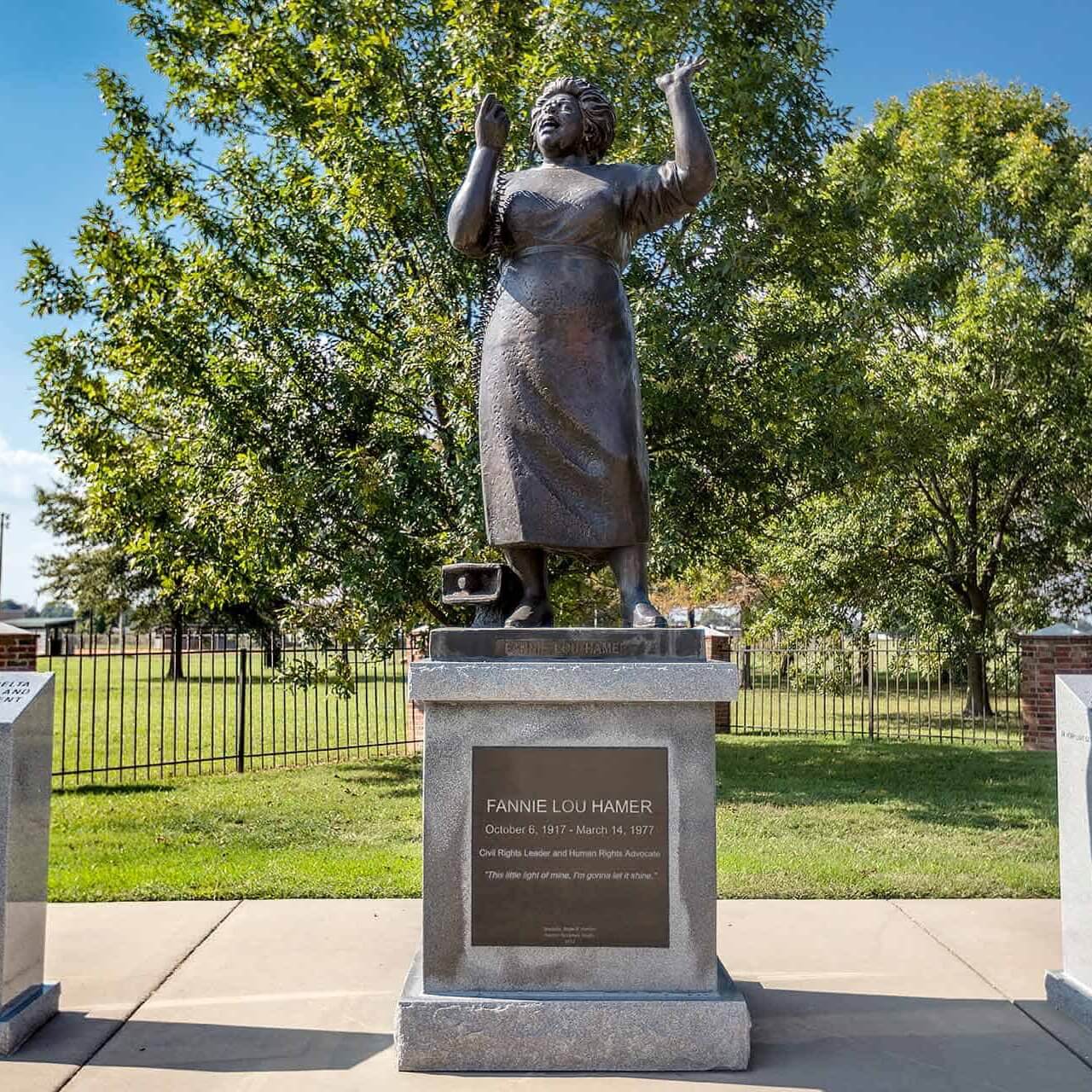 A bronze statue of a woman stands proudly on a pedestal with an engraved plaque, guiding visitors along Mississippi's Freedom Trail. In the background, trees sway gently behind a rustic fence.