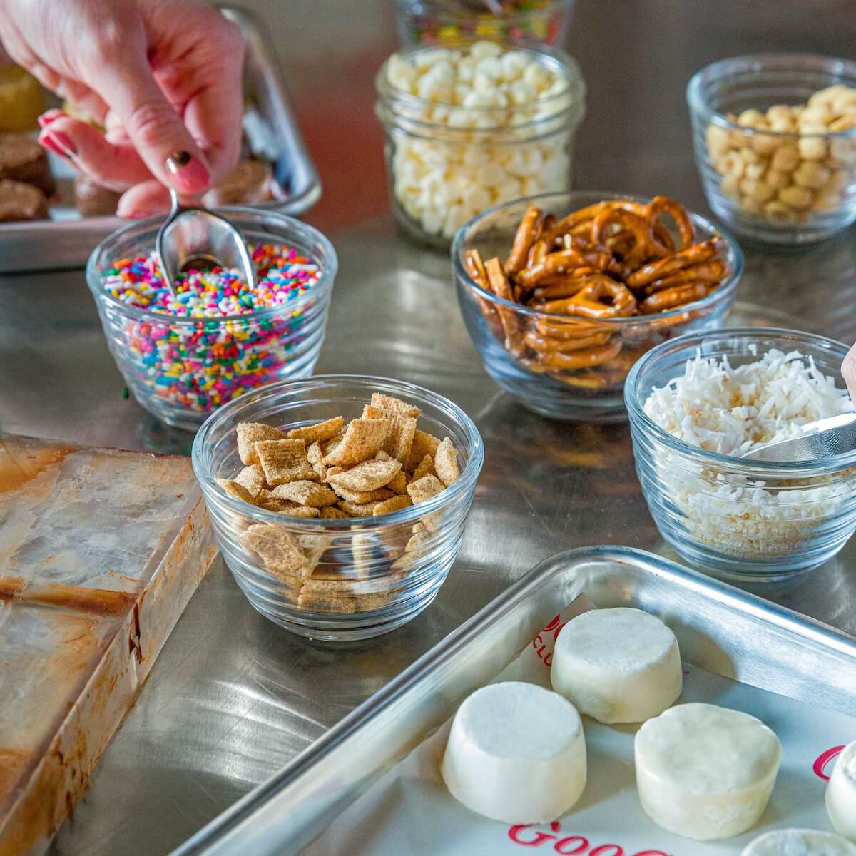 A selection of toppings in glass bowls, including colorful sprinkles, pretzels, cereal squares, and coconut shreds, with a hand holding a spoon over the sprinkles—perfect for a day of family fun in Nashville.