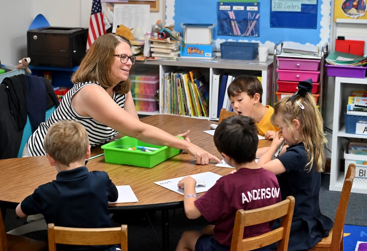 A teacher sits at a table with four children, diligently helping them with their work in the classroom. 