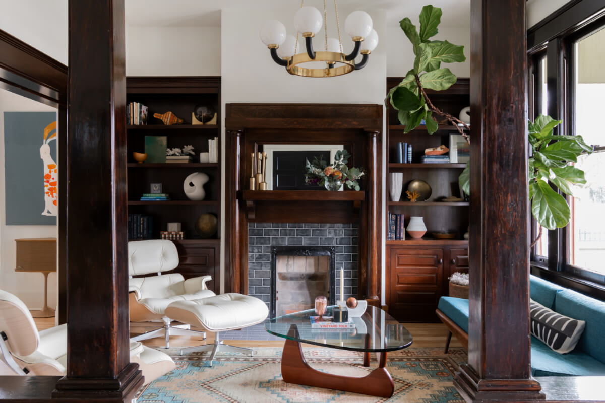 A Historic Nashville Home with STUNNING Colorful Accents! - 2 A cozy living room by Sara Ray features dark wood accents, a brick fireplace, a white lounge chair, and a glass coffee table. A large green plant adds freshness, while shelves showcase books and decorative items.