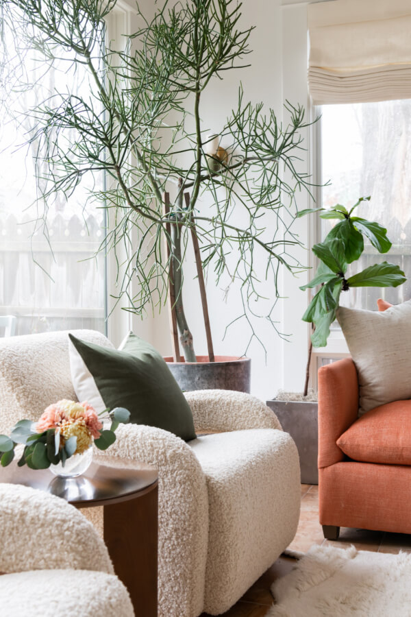 A Historic Nashville Home with STUNNING Colorful Accents! - 11 Sara Ray's design features a cozy living room with a textured armchair, orange sofa, and large potted plants. A small wooden side table holds a floral arrangement, perfectly complementing the white-blinded window.