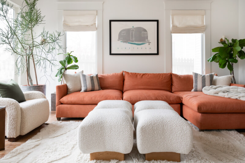 A Historic Nashville Home with STUNNING Colorful Accents! - 10 This living room features an orange sectional sofa, two white ottomans, and a cozy armchair. Large potted plants add greenery, while a framed artwork on the wall completes this inviting home feature.