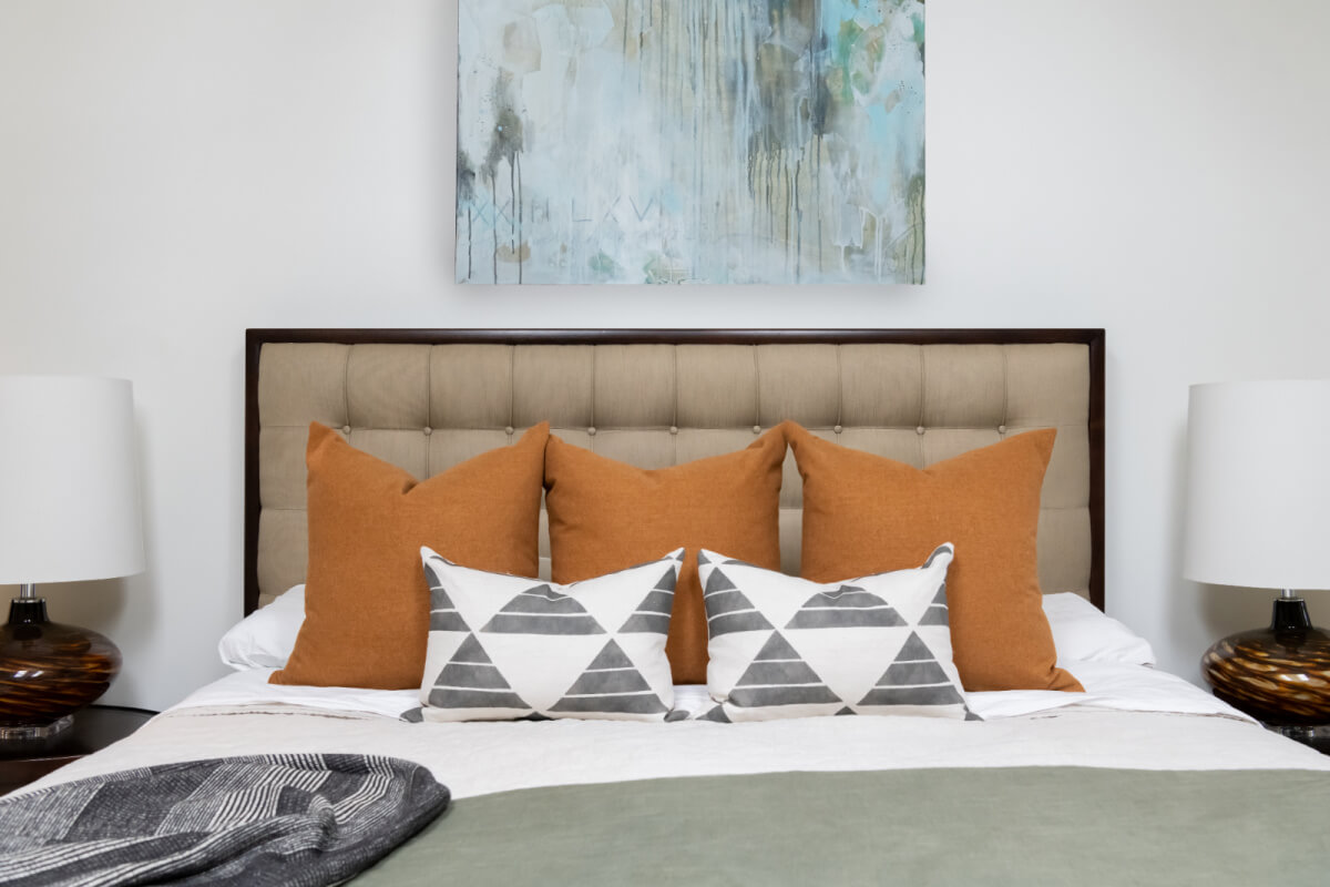 A Historic Nashville Home with STUNNING Colorful Accents! - 13 A neatly made bed with brown and patterned pillows against a tan headboard, flanked by two lamps. A modern abstract painting hangs on the wall above.