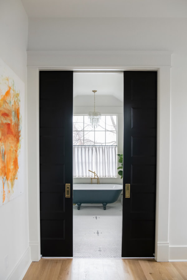 A Historic Nashville Home with STUNNING Colorful Accents! - 14 View of a bathroom designed by Sara Ray, featuring black framed sliding doors, a vintage tub, a chandelier, and a window with sheer curtains.