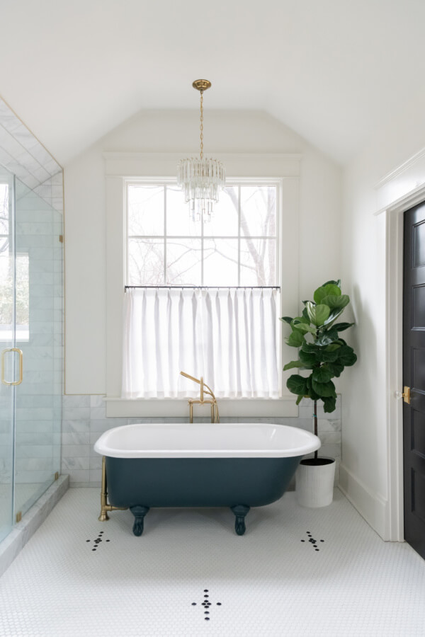 A Historic Nashville Home with STUNNING Colorful Accents! - 15 This bathroom features a freestanding clawfoot tub and a leafy plant. A chandelier adds elegance, while a window with white curtains lets in natural light. The floor is tiled in an intricate white pattern.