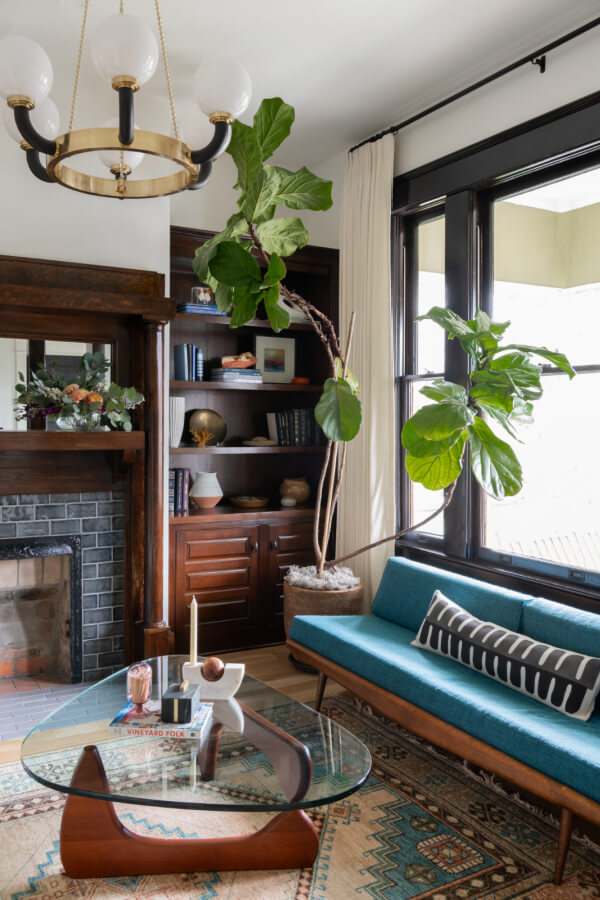 A Historic Nashville Home with STUNNING Colorful Accents! - 3 A cozy living room features a teal sofa, glass coffee table, and large potted plant. Bookshelves and a fireplace grace the background, while a black chandelier adds elegance to the ceiling.