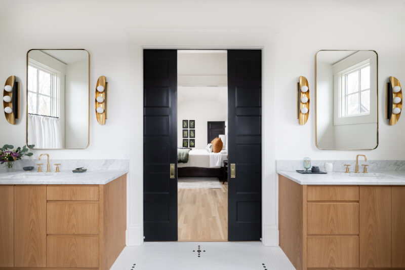 A Historic Nashville Home with STUNNING Colorful Accents! - 16 Modern bathroom with dual wooden vanities, gold fixtures, and large mirrors. Black double doors feature access to a bedroom where sunlight streams through windows, illuminating the bed.