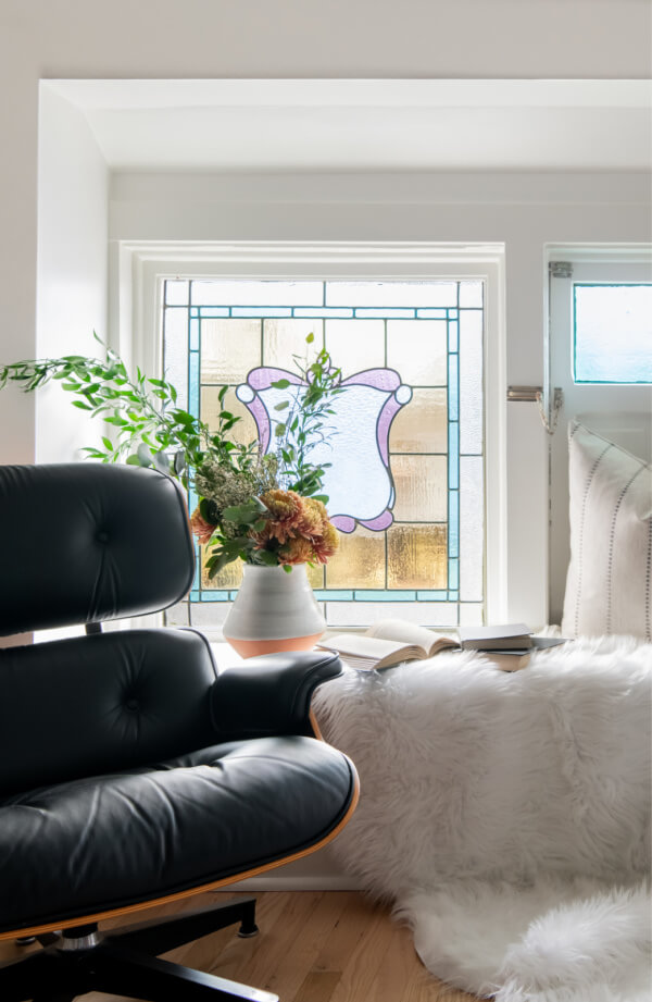 A Historic Nashville Home with STUNNING Colorful Accents! - 19 A cozy corner features a black chair, fuzzy white rug, and a window with stained glass. A vase with flowers and an open book are artfully placed on the rug.