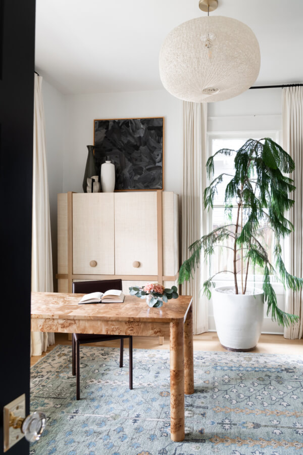 A Historic Nashville Home with STUNNING Colorful Accents! - 17 A minimalist home office features a wooden desk, an open book, a tall plant in a white pot, and a large cabinet. A textured light fixture adds charm as natural light filters through white curtains.