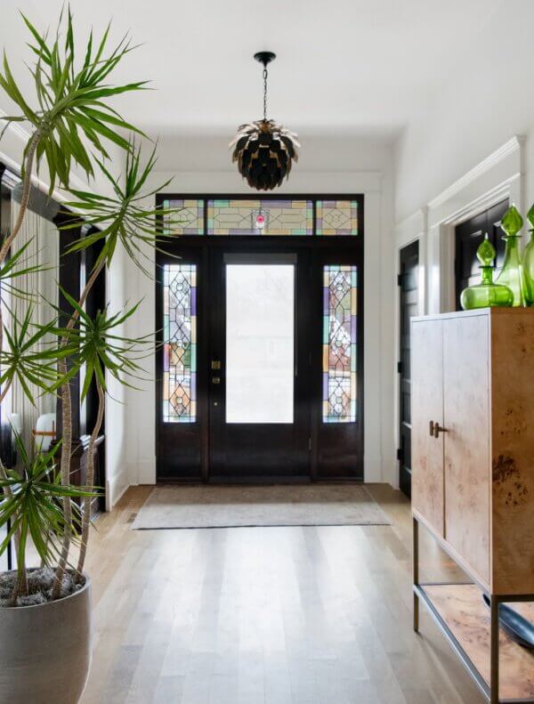A Historic Nashville Home with STUNNING Colorful Accents! - 1 A hallmark of Sara Ray, this hallway boasts elegant wood flooring, a large potted plant, and a cabinet adorned with green glass decor. The space is beautifully completed by a stunning stained glass front door—a truly captivating home feature.