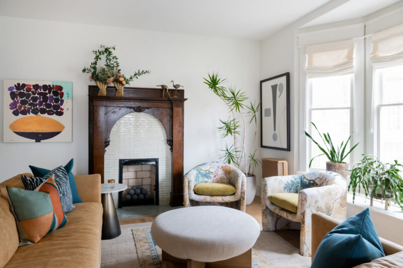 A Historic Nashville Home with STUNNING Colorful Accents! - 4 A cozy living room features a tan sofa, patterned armchairs, and a large circular ottoman. Artwork and plants adorn the space, while a decorative fireplace and large windows flood the home with natural light.