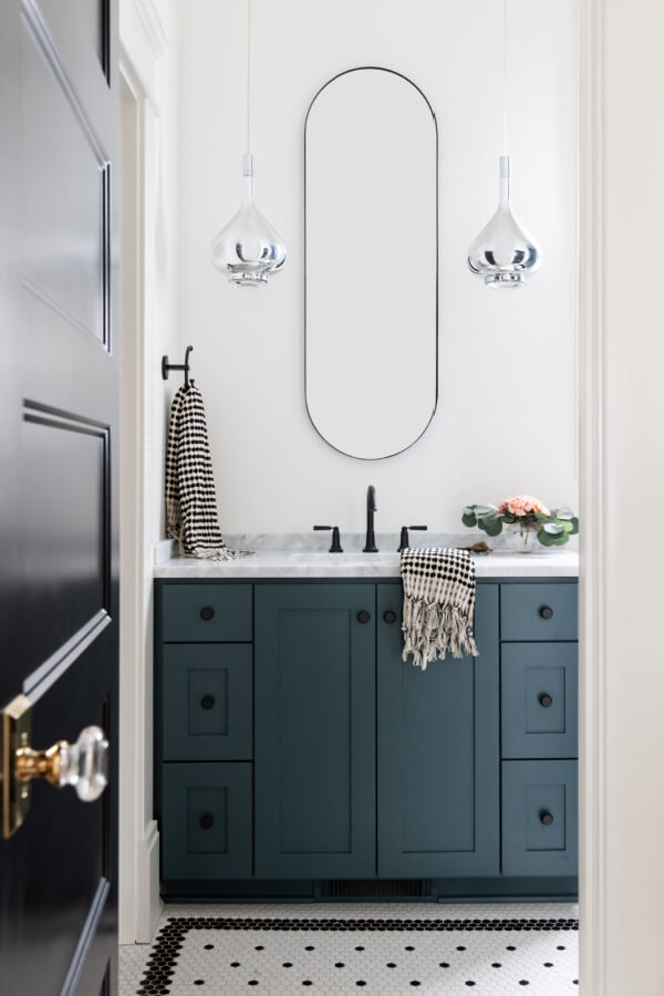 A Historic Nashville Home with STUNNING Colorful Accents! - 12 A stunning bathroom by Sara Ray features a black and white tiled floor, a dark green vanity, an oval mirror, silver pendant lights, and a checked towel. This elegant home feature perfectly balances classic charm with contemporary style.