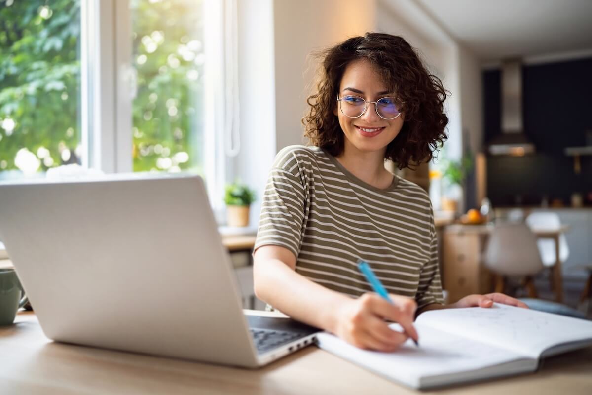 A person with curly hair and glasses writes in a notebook while sitting at a desk with an open laptop in a bright, modern room, eager to learn from experts after enrolling in the best masterclasses.