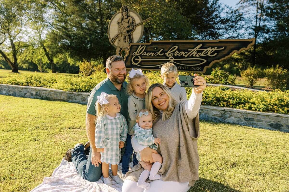 A family of five captures a joyful travel selfie on a cozy blanket, perfectly framed by a welcoming park sign during their Tennessee vacation.