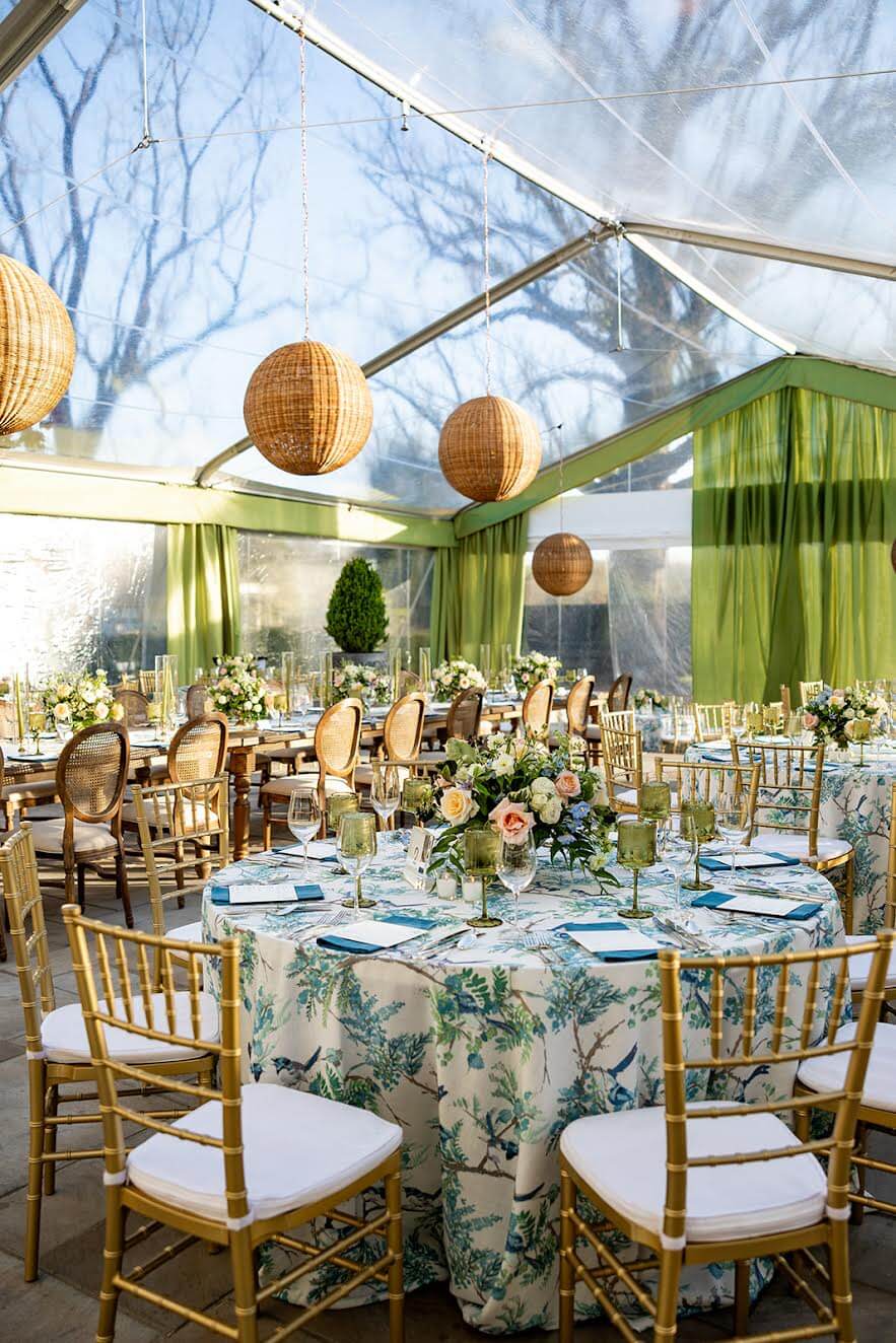 Round tables with floral tablecloths and gold chairs, arranged by the best event planners, are set for an event under a clear tent with hanging wicker lanterns and green drapes.