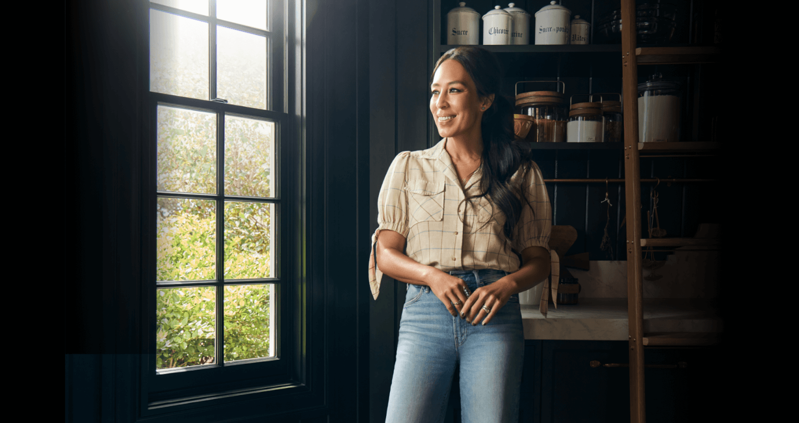 A person stands in a kitchen, wearing a checkered shirt and jeans, near a window with natural light streaming in. 
