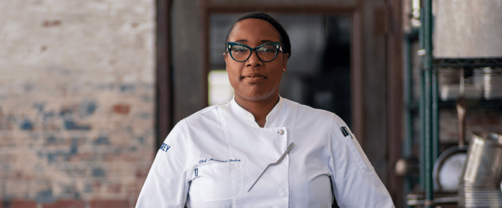 A Southern chef, wearing glasses and a pristine white coat, stands confidently in the kitchen.