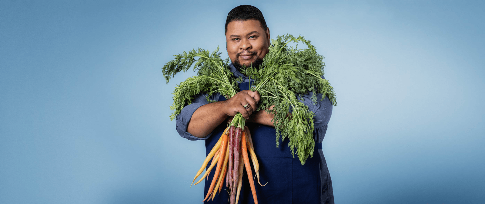 Against a blue background, a person in a blue apron proudly holds a bunch of colorful carrots with green tops, embodying the spirit of Southern Masters learning.