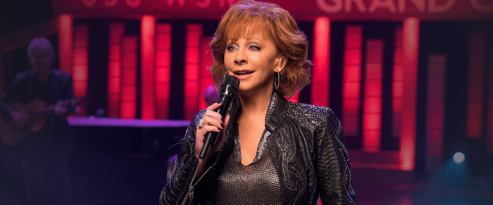 Reba grips a microphone on stage amidst a blurred backdrop of twinkling lights and musicians.