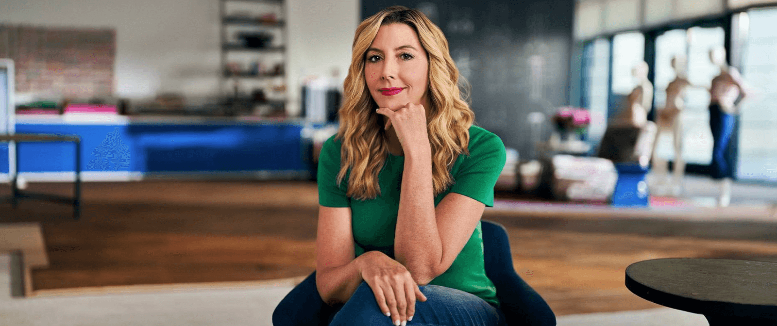 A person with long blonde hair sits in a modern room. They wear a green shirt and blue jeans, their hand resting thoughtfully on their chin.