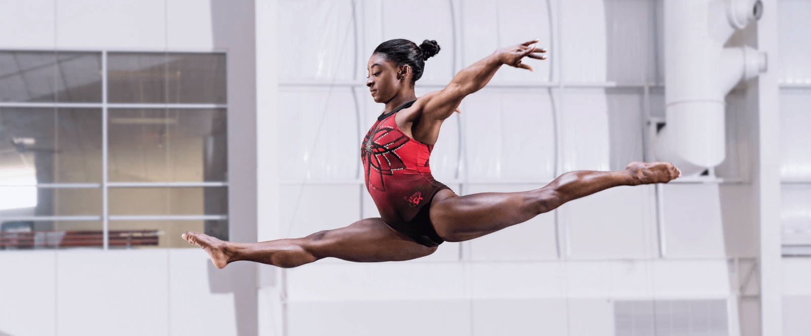 A gymnast performs a mid-air split leap in a gymnasium, gracefully showcasing her skills in a red and black leotard.