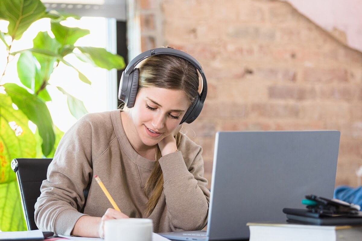 A person discovers their hidden genius while wearing headphones, jotting notes in a notebook, and gazing at an open laptop on the desk, accompanied by a plant in the background.