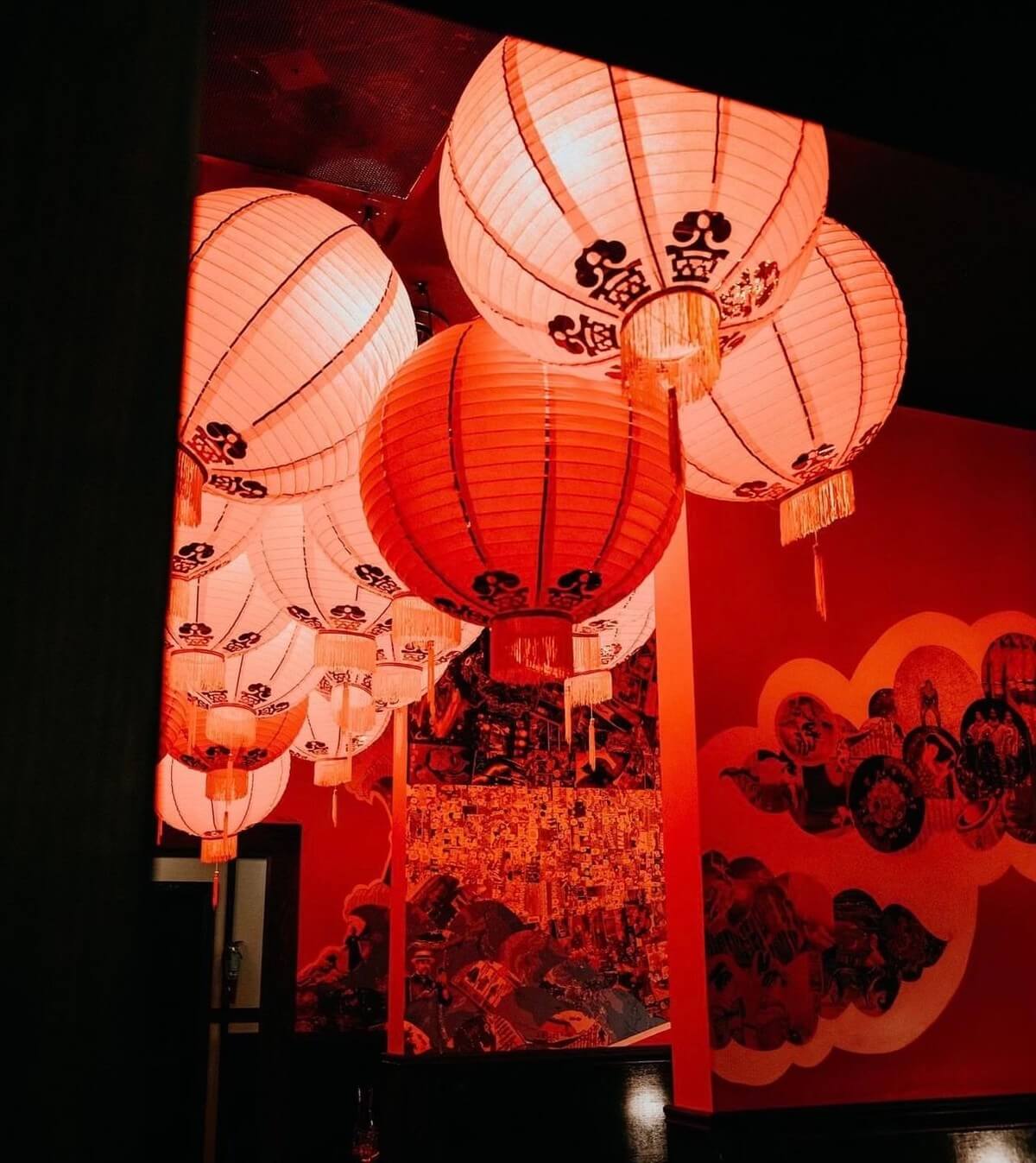 Red Chinese lanterns with black decorative patterns hang from a ceiling in a dimly lit room, adorned with red walls and decorative murals.