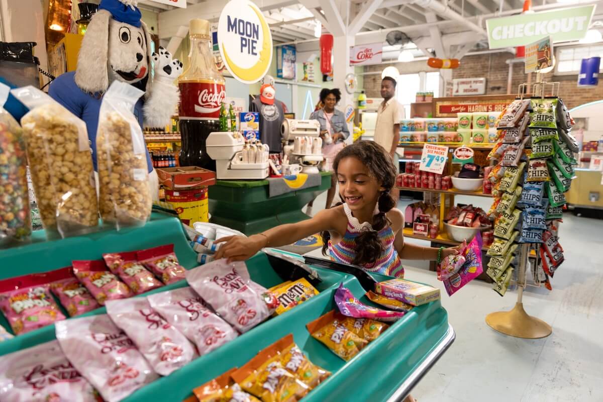 In a vibrant dress, a girl reaches for candy at a retro-style store in Hattiesburg, MS. Various snacks and vintage signs create an inviting atmosphere.