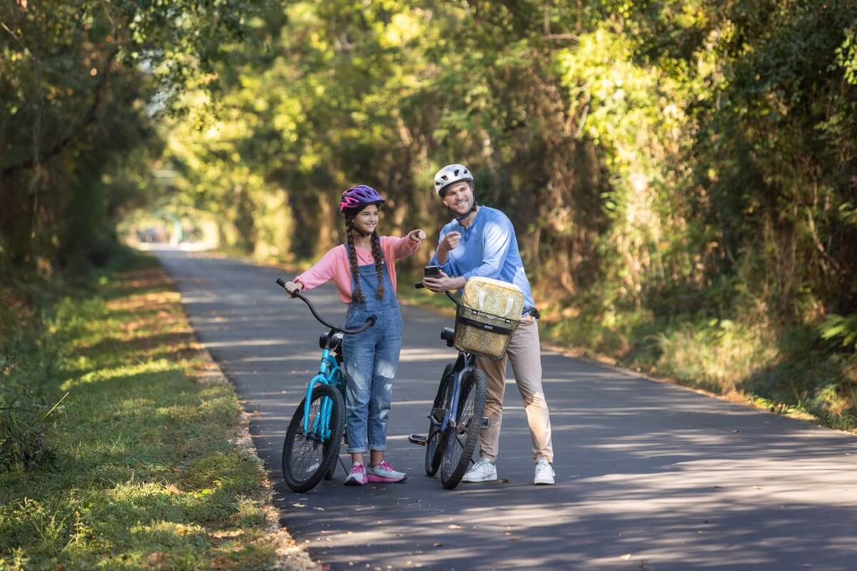 A man and a girl, both wearing helmets, stand beside their bicycles on a sunny, tree-lined path in Hattiesburg, MS. They appear to be smiling and pointing at something.