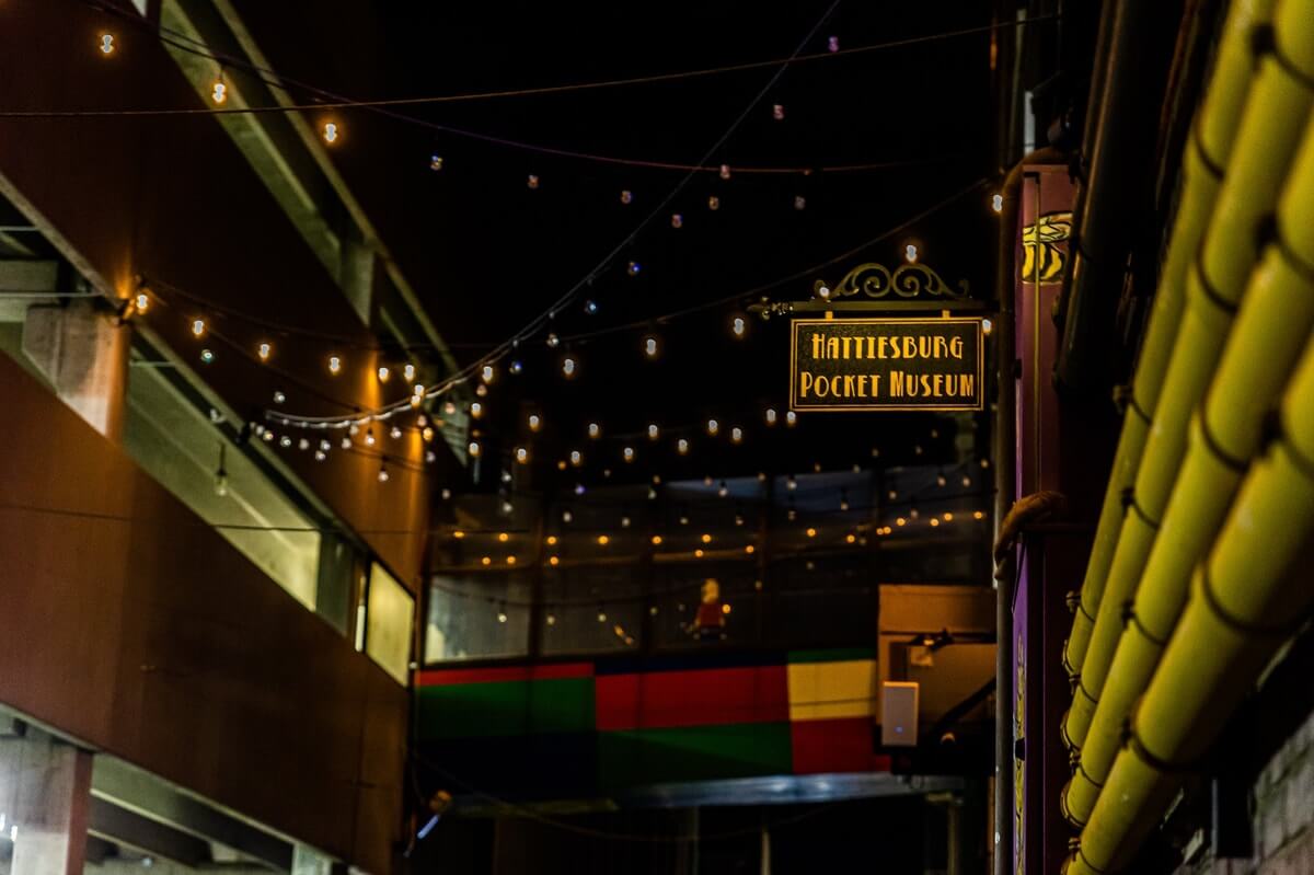 Night scene of an alleyway with string lights. A sign reads "Hattiesburg Pocket Museum." A colorful building is in the background.