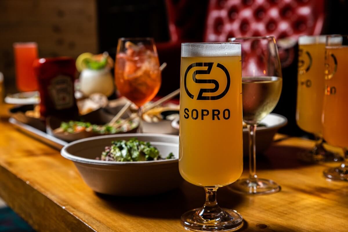 A glass of beer with "SOPRO" branding is on a wooden table, surrounded by various drinks and dishes. Background features a blurred, plush red chair.