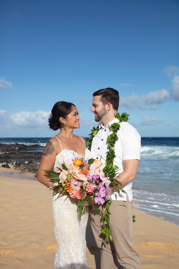 A couple in wedding attire stands on a beach, the woman holding a bouquet of colorful flowers. Under the clear blue sky, Lennie and her husband gaze lovingly at each other.