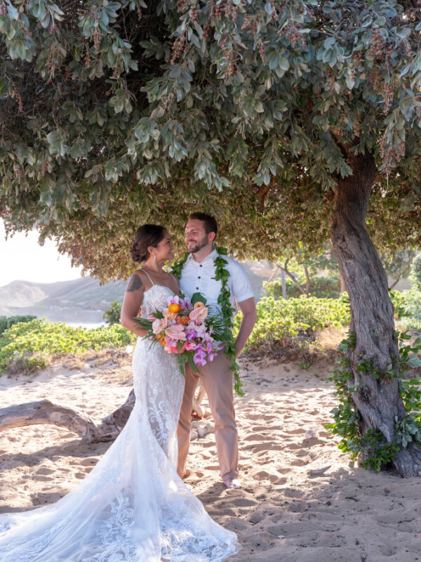 A couple in wedding attire stands on a sandy beach under a tree, holding a colorful bouquet.