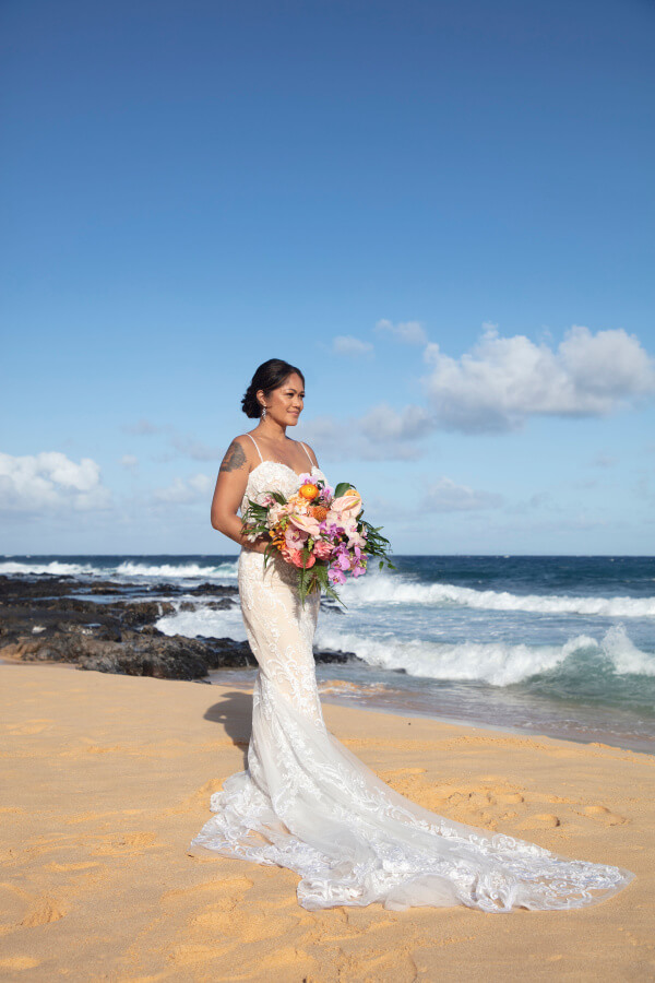 A woman in a long white wedding dress holds a bouquet while standing on a sandy beach with the ocean in the background, capturing the perfect moment of her dream wedding.