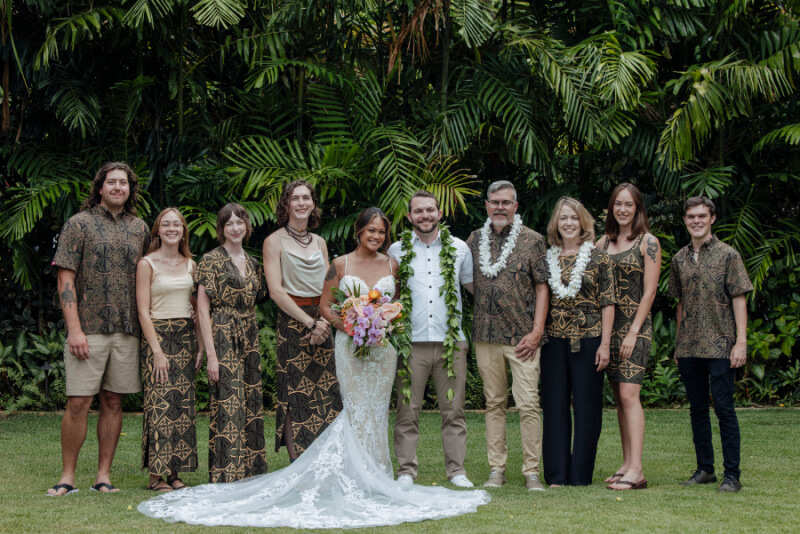 A wedding party stands together outdoors in front of Lennie's lush greenery. The group, including the bride, groom, and guests in patterned attire, radiates joy. The bride holds a bouquet.