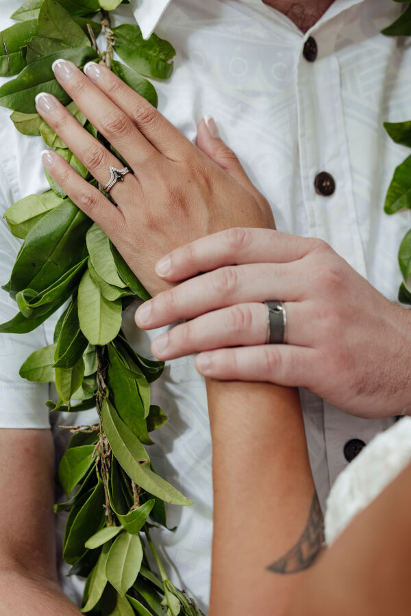 Close-up of a couple holding hands, one adorned with a leafy lei and white shirt, both showcasing shining wedding rings.