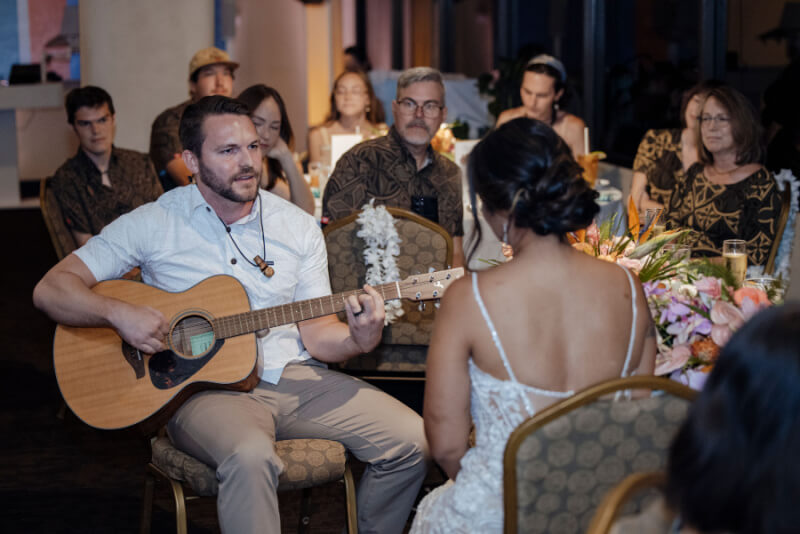 William strums an acoustic guitar, while guests in the background watch attentively.