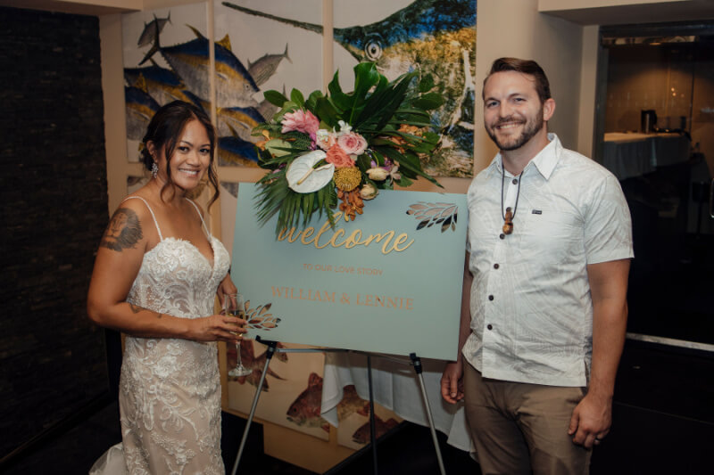 A bride and groom stand beside a beautifully decorated welcome sign at their wedding. The sign reads "Welcome to our love story - William & Lennie," with stunning floral decor. 
