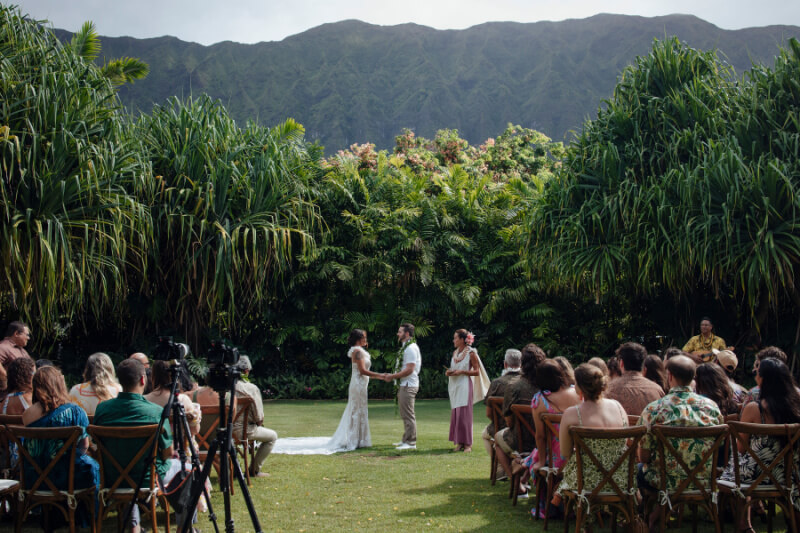 Lennie and William stand at the altar during a breathtaking outdoor wedding ceremony, surrounded by guests seated on wooden chairs, with lush greenery and majestic mountains providing a stunning backdrop.