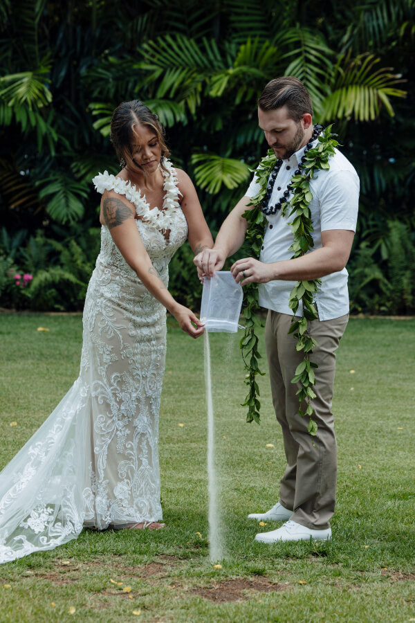 Lennie and her partner elegantly perform a sand pouring ceremony during their wedding, surrounded by lush greenery in the picturesque outdoor setting.