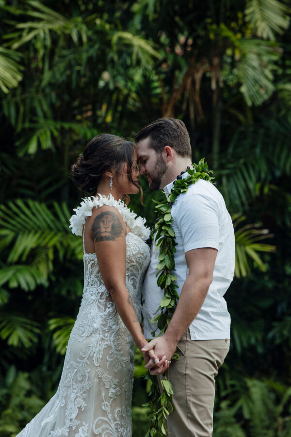 Lennie and her partner, dressed in elegant wedding attire, stand facing each other, holding hands amid the lush greenery. Each wears a lei, symbolizing their love and connection in this enchanting setting.