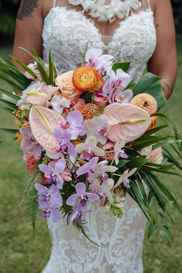 A person in a white lace wedding dress holds a bouquet of pink and orange flowers, including orchids, roses, anthuriums, and greenery, against a grassy background.