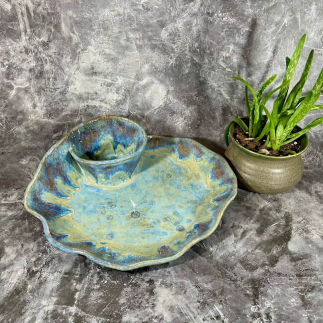 A blue-green ceramic chip and dip platter with a ruffled edge sits beside a small green plant in a brown pot, adding charm on a textured gray background.