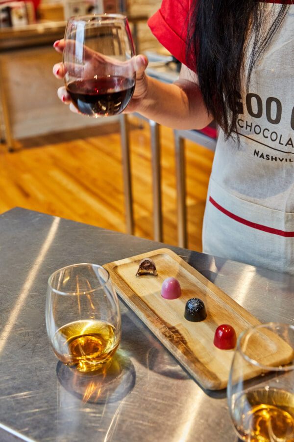 In a Nashville kitchen, someone in a red shirt and apron holds a glass of red wine by a wooden platter with four chocolates. Nearby, another glass of amber liquid adds to the warmth of the scene, perfect for winding down after adult education classes.