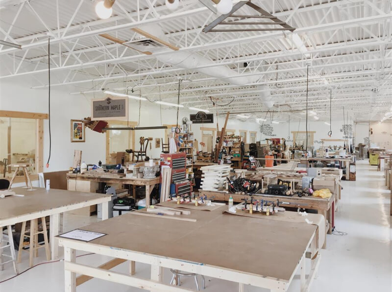  Large tables, tools, supplies, and machinery are thoughtfully arranged throughout a workshop space. Bright lighting and high ceilings complete the inviting and open atmosphere for creative exploration.