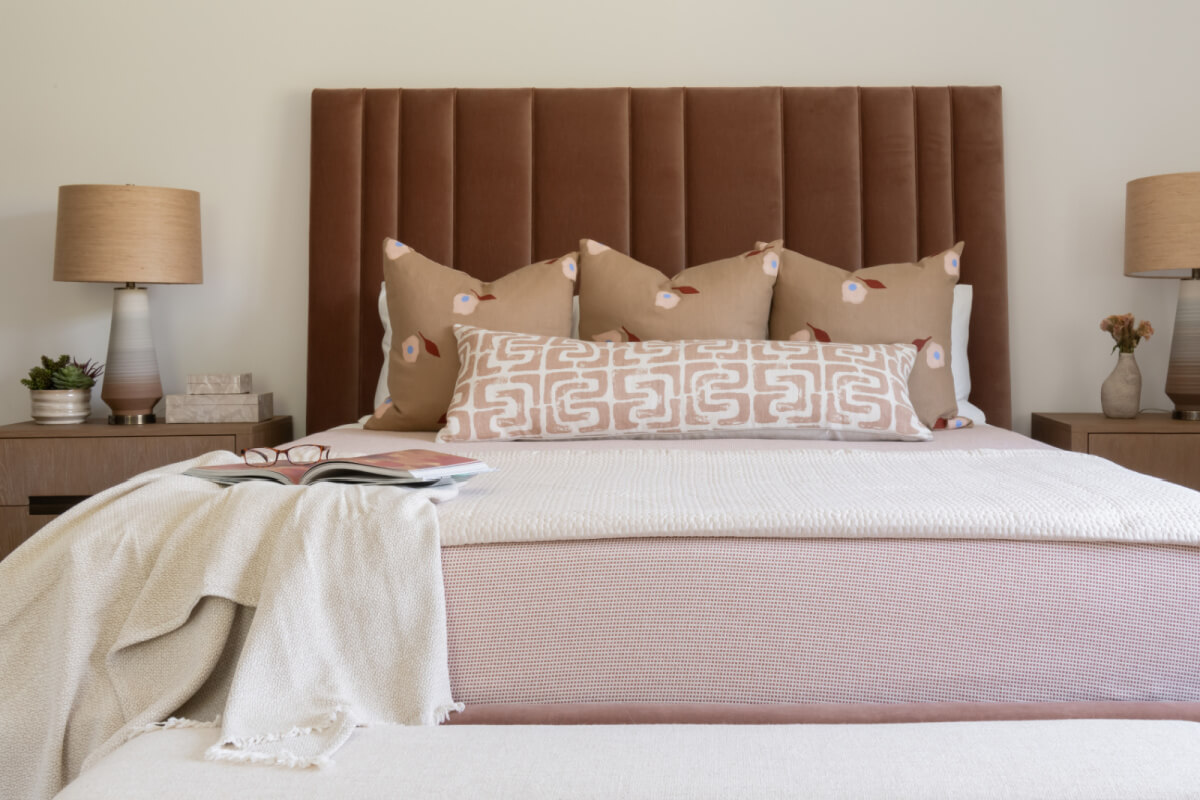Cozy bedroom with a textured brown headboard, neutral bedding, and decorative pillows reflecting 2025 interior design trends. Two bedside tables with lamps flank the bed, where a book and throw blanket add comfort.