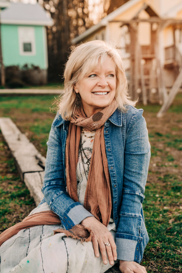 Susan Binkley, with her blonde hair cascading over a denim jacket and scarf, sits outdoors, her face beaming with a warm smile. In the background, there's a grassy area with a wooden beam and quaint houses nestled under the clear sky.