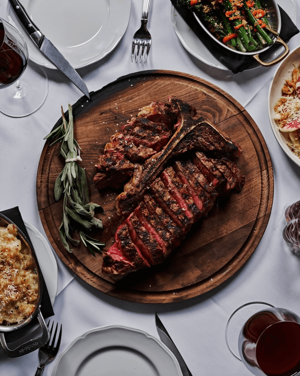 A sliced, grilled steak on a wooden platter with rosemary, surrounded by side dishes, glasses of red wine, and plates on a white tablecloth—a must-try dish at one of Atlanta's new restaurants this year.