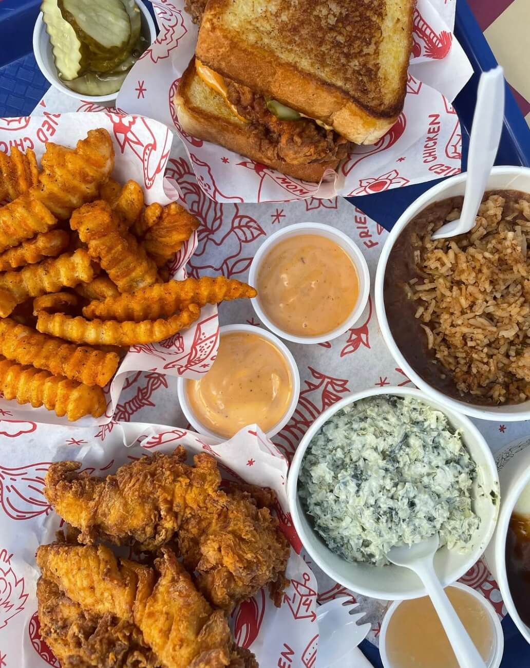 Discover a delicious meal on a tray at new Atlanta restaurants featuring a fried chicken sandwich, crinkle-cut fries, fried chicken pieces, dipping sauces, rice, and a bowl of spinach dip.