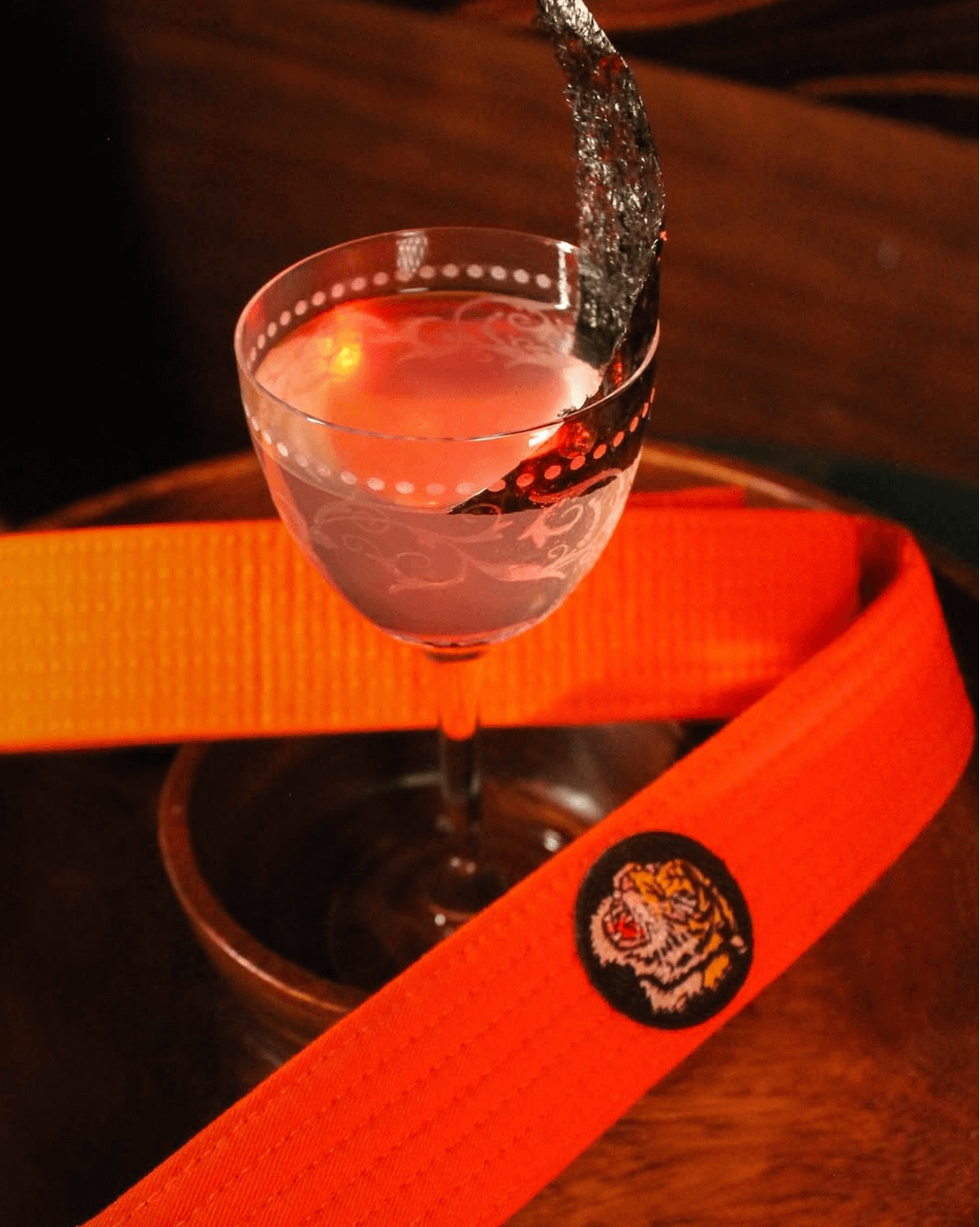 A cocktail glass with a decorative rim, wrapped in a strip, sits next to an orange belt featuring a tiger emblem on a wooden surface. Discover this innovative presentation at one of the new restaurants you must try this year.